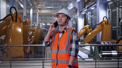side view of asian male engineer with safety helmet talking on smartphone while standing with industrial robotic arms working