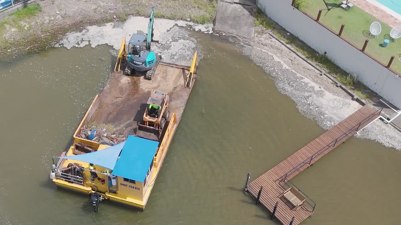 Aerial footage of a barge carrying an excavator, positioned near a residential dock and retaining wall.