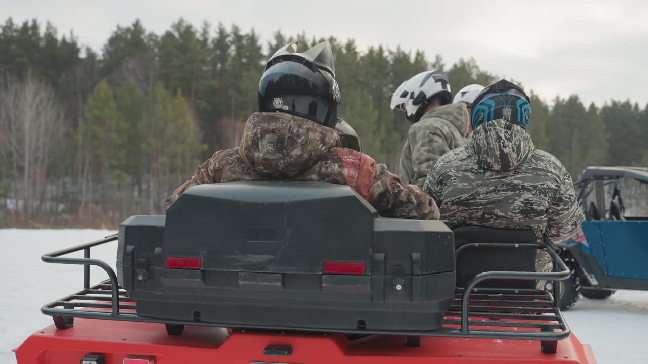Due passeggeri sul sedile posteriore dell'ATV in una foresta innevata, giacche mimetiche e caschi, un ATV utility rosso che attraversa un lago ghiacciato con un UTV che passa accanto, pneumatici robusti che sollevano la neve, luce calma e nuvolosa, attesa.