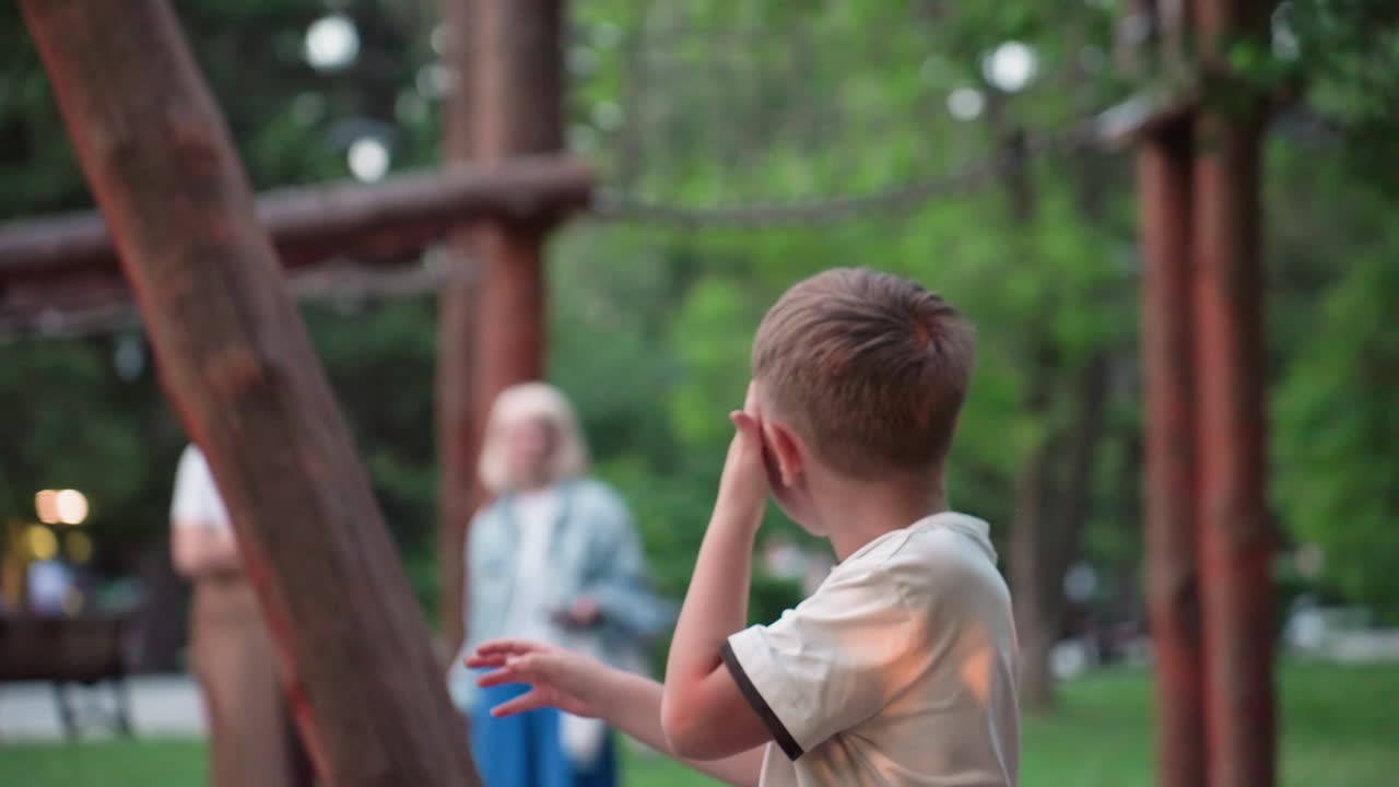 back view of playful boy pushing his friend on wooden chain swing in green park at dusk with warm sunset glow capturing joyful motion blur of summer trees and equipment during carefree outdoor
