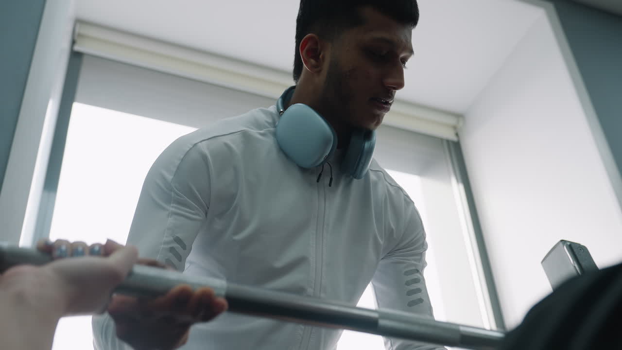 close up of gym instructor in white jacket with headphones around neck guiding student during barbell lift, focused expression and supportive hand positioning in bright indoor fitness training space