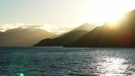 light rays stream across tops of mountain peaks casting beams over lake wakatipu