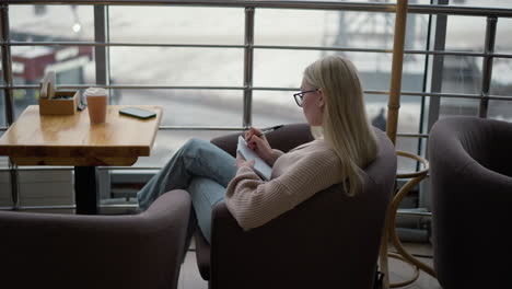vista lateral de una mujer sentada en una mesa con un libro y una taza de café, escribiendo pensativamente en un cuaderno, mostrando una atmósfera acogedora y enfocada, con un fondo suave de una cafetería o espacio de oficina