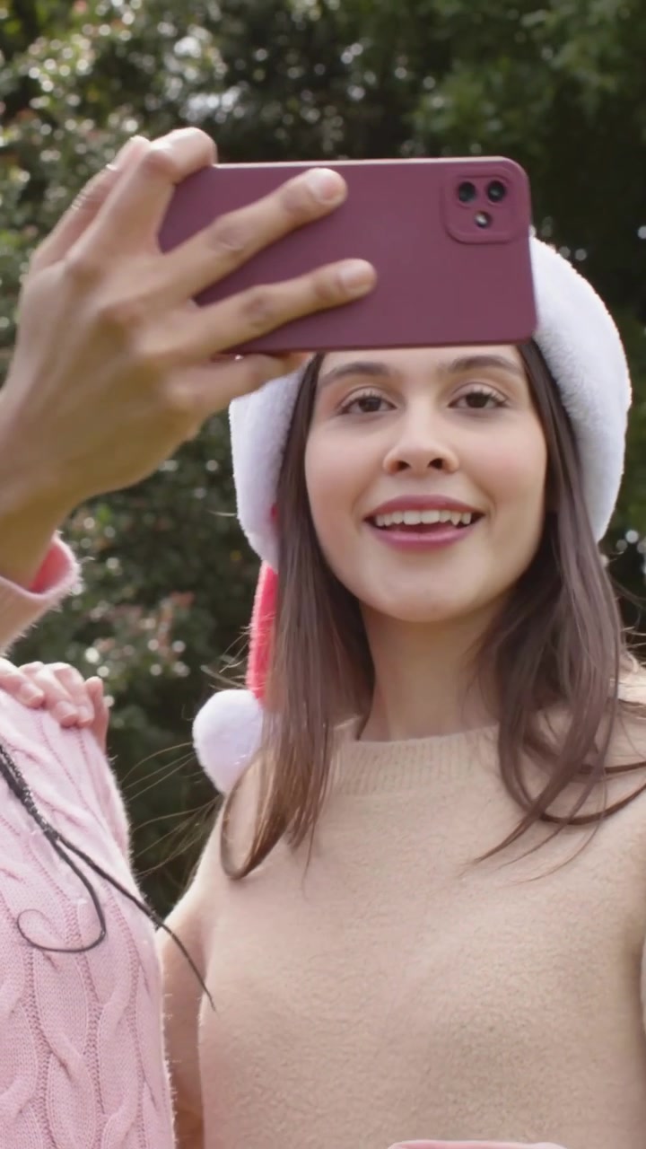 Video verticale: Donne sorridenti con cappelli da Babbo Natale che posano in un parco, quella a sinistra alza un telefono rosa per catturare la gioia