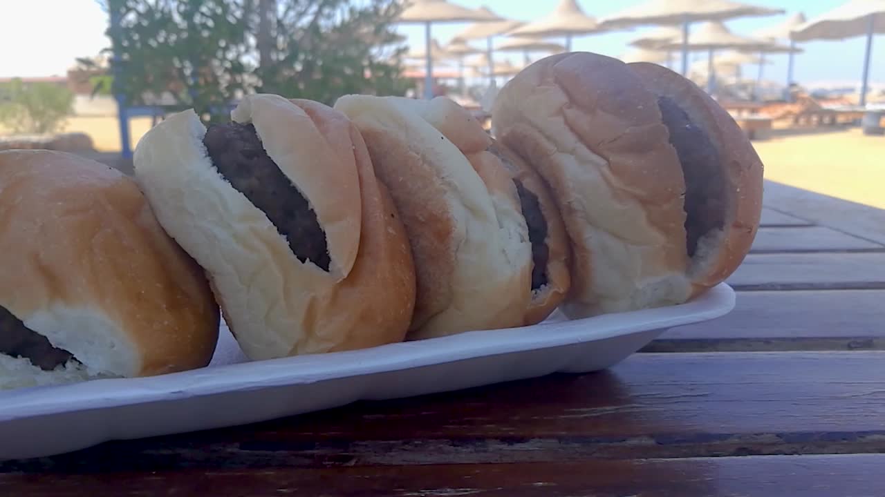 백그라운드에서 비치 파라솔이 있는 나무 테이블에 앉아 있는 스티로폼 접시에 햄버거 4개