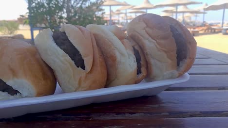 Four-burgers-on-styrofoam-plate-sitting-on-wooden-table-with-beach-umbrellas-in-the-background