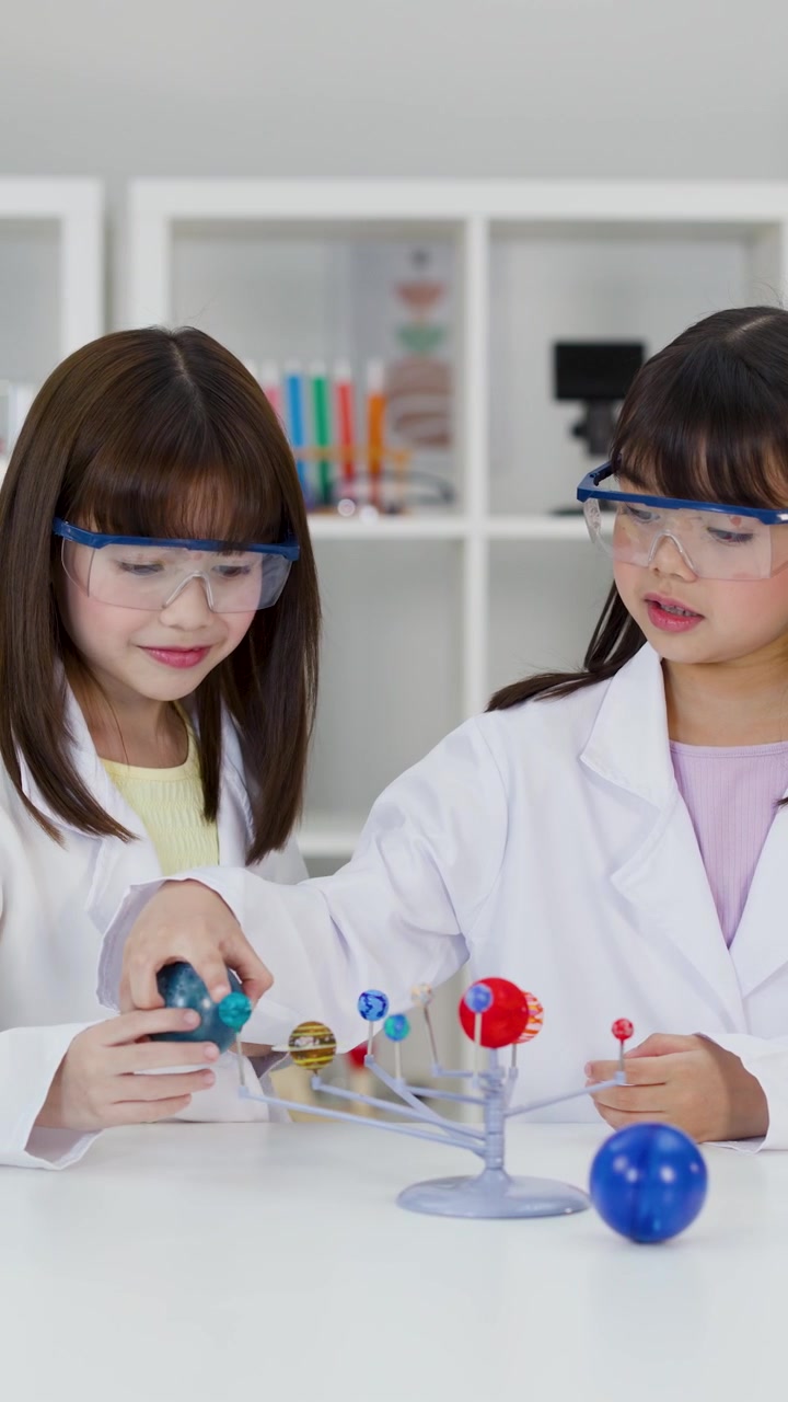 Dua gadis mengenakan jas lab dan kacamata pelindung memeriksa model planet di ruang kelas yang terang.