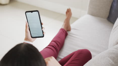 Biracial-woman-sitting-on-sofa-using-smartphone-with-copy-space-on-screen,-slow-motion