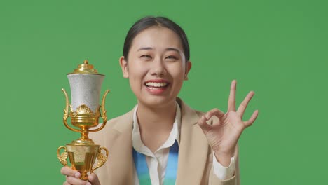 close up of asian business woman in a suit with a gold medal and trophy showing okay gesture and smiling to camera as the first winner on green screen background in the studio