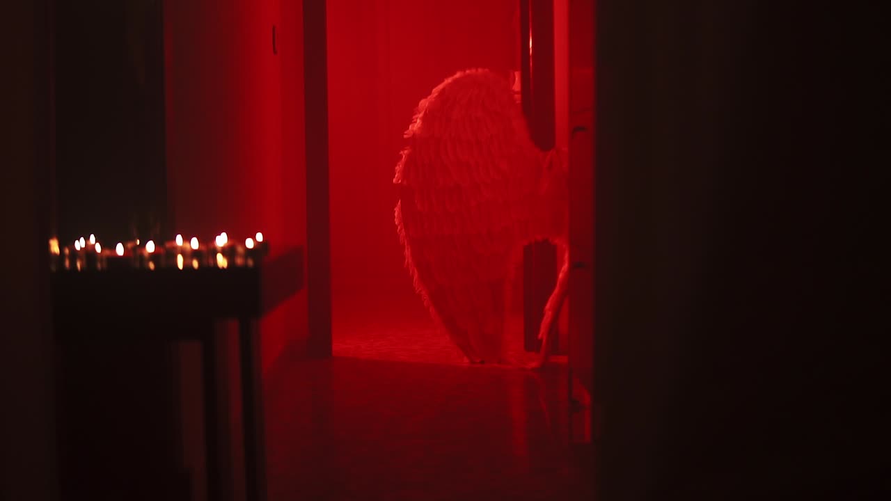 Ripresa cinematografica in slow motion di un palco con luci d'ambiente rosse e candele su un tavolo, ali piumate bianche sullo sfondo.