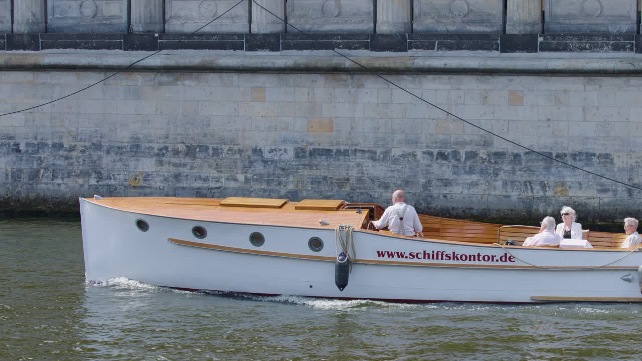 A small white tour boat with passengers moves steadily along a riverside wall in Berlin under daylight, captured in a smooth lateral tracking shot