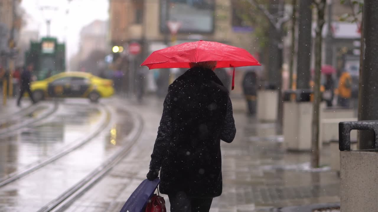 Woman holding red umbrella walks in city as snow falls in slow motion