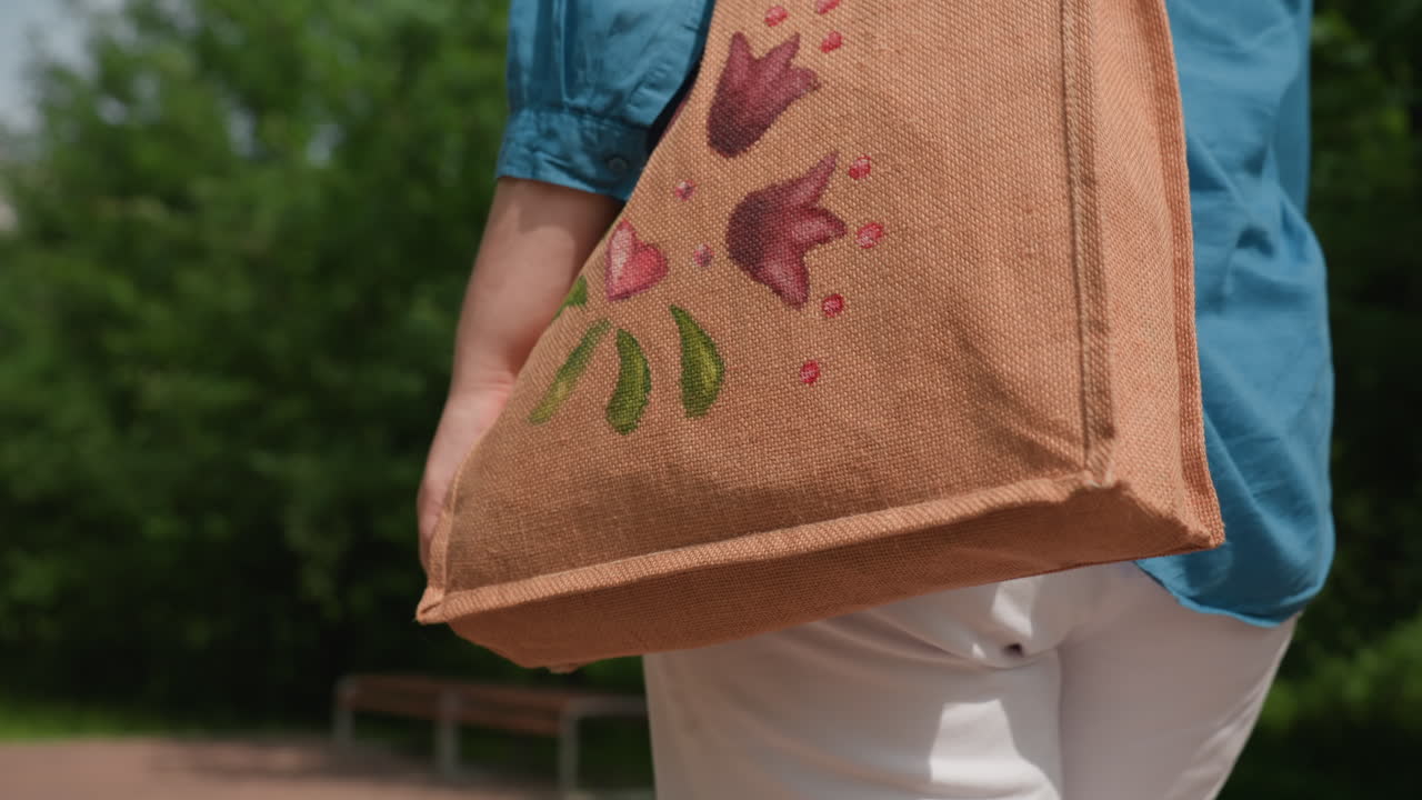 푸른 셔츠와 흰 바지를 입고 화려한 꽃무늬가 수놓아진 갈색 가방을 멘 여성이 초록 잎사귀로 둘러싸인 화창한 공원 길을 따라 걷는 뒷모습 클로즈업 뷰