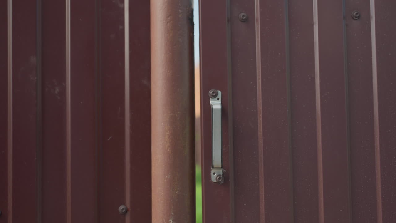 Rear view of person in denim jacket opening metal gate with hand on handle, partially blurred backpack in foreground, captured during bright sunny day with strong shadows and outdoor ambiance