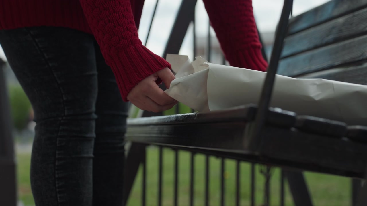 Close up of woman in red sweater and dark jeans approaching then standing next to outdoor swing. Woman interacts with wrapped items on bench seat as soft daylight illuminates grassy park