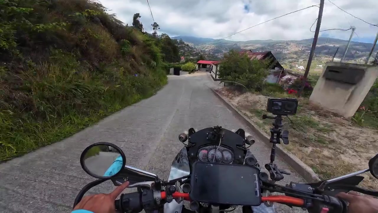 Sepeda motor melaju di jalan pegunungan yang berkelok-kelok. Dikelilingi oleh perbukitan hijau yang rimbun, vegetasi di tepi jalan, dan langit berawan, pemandangan ini menangkap esensi perjalanan pedesaan yang indah.