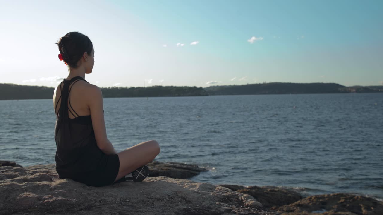 mujer joven sentada con las piernas cruzadas sobre rocas junto al agua durante la puesta de sol