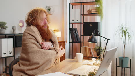 sick businessman shivering from the cold wrapped in blanket sneezes works on laptop at office