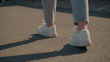 close-up back view of individual walking on sunlit tarred road, wearing white sneakers and jeans, shadows are cast on ground, capturing casual movement, outdoor setting