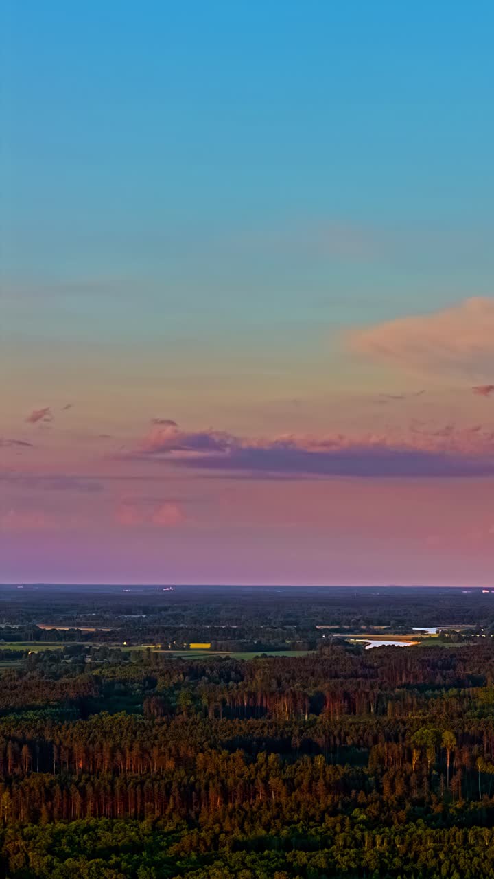 Vivaci nuvole rosa fluttuanti sopra una vasta foresta verde e un paesaggio lacustre nella campagna lettone durante il tramonto - timelapse verticale cinematografico con drone