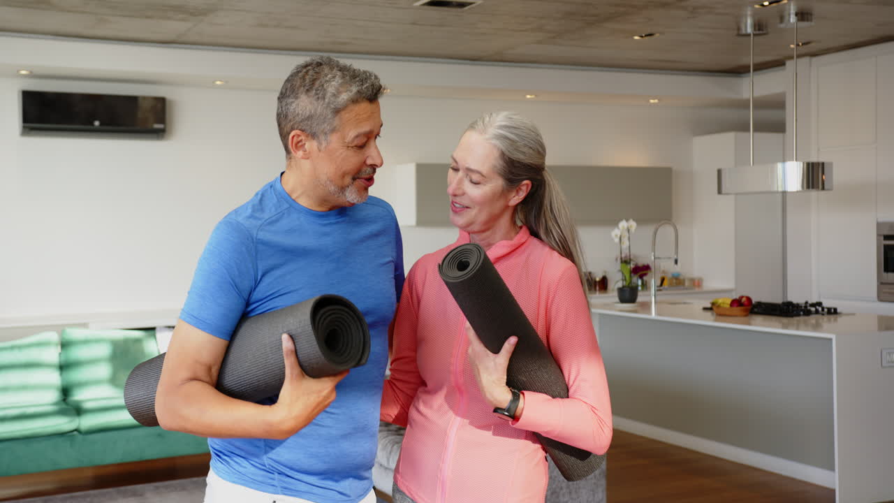 Holding yoga mats, smiling multiracial senior couple preparing for home workout in modern kitchen