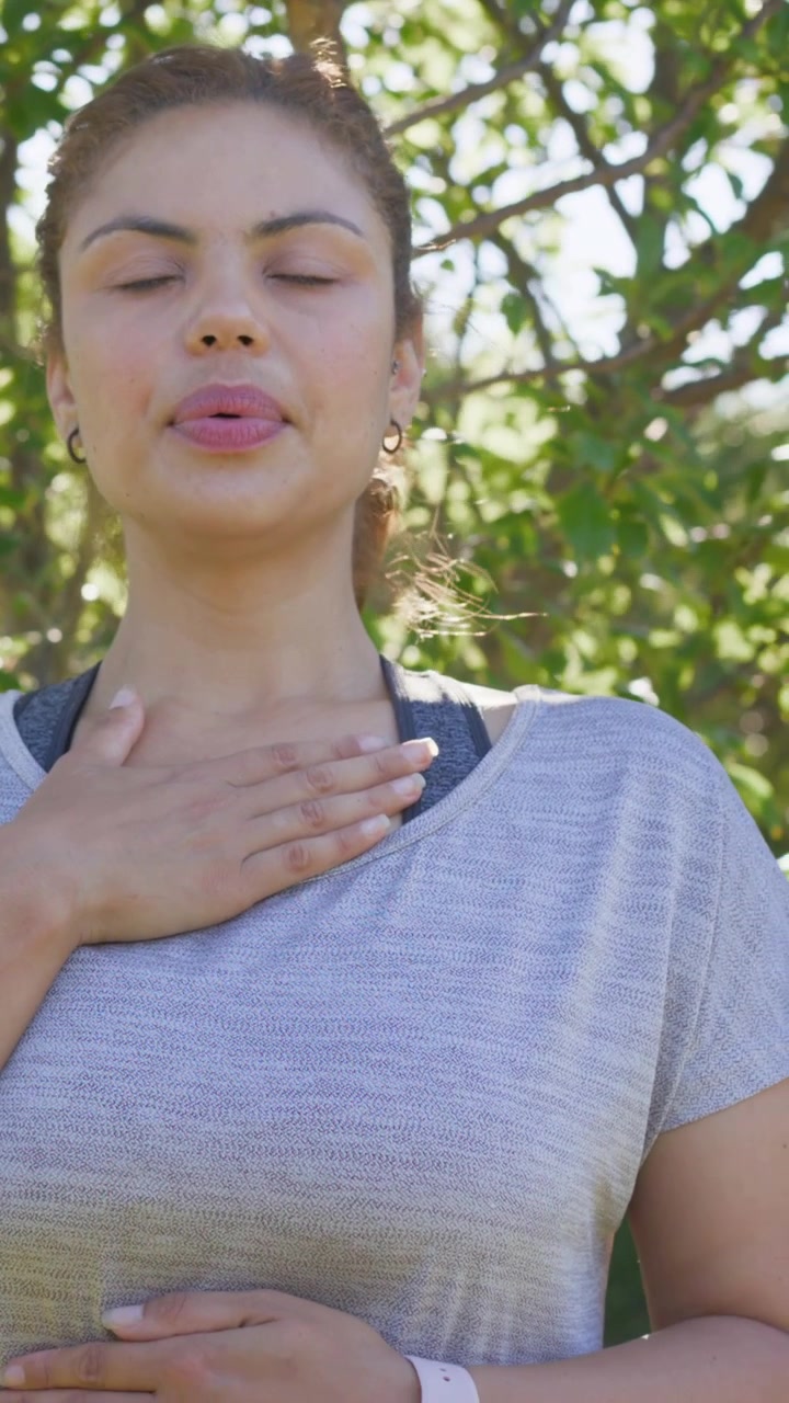 Vídeo vertical: Mulher de blusa respirando com os olhos fechados e as mãos no peito em um parque, relaxando com um relógio.