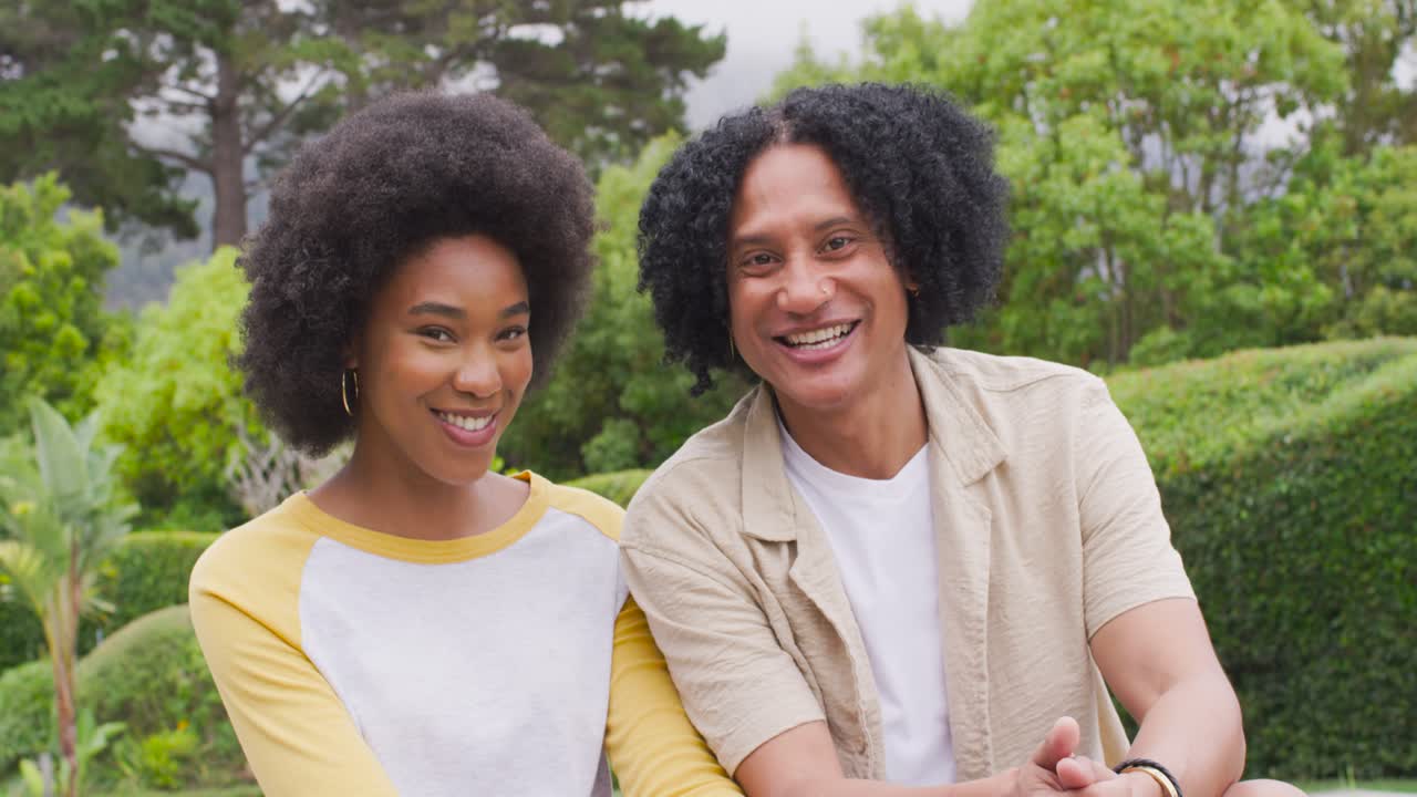 Casal afro-americano sentado em um banco no jardim, o homem iniciando uma conversa divertida e fazendo a mulher sorrir.