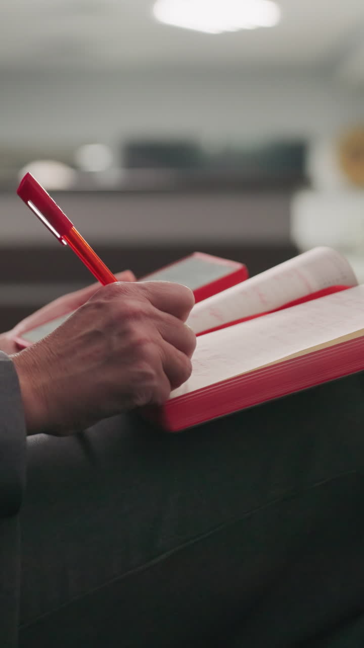 Businesswoman sitting in modern office, looking at red notebook and mobile phone, multitasking, focused on task, reading, corporate workspace, gray suit, productivity, concentration