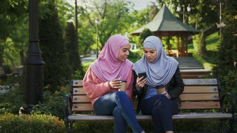 two dark-skinned muslim women in colorful hijabs. they laughing, talking, looking at screen of smartphone. sitting on bench in park. сlose up