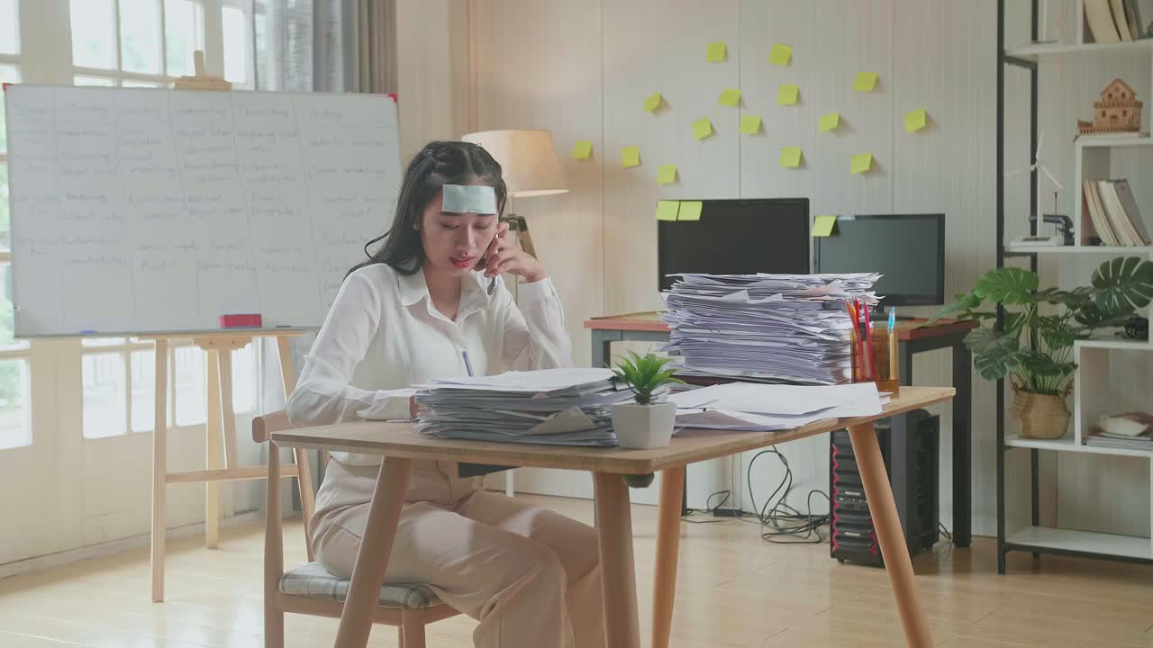 병든 아시아 여성이 사무실에서 문서와 함께 열심히 일하면서 전화로 이야기하고 메모를 취합니다.