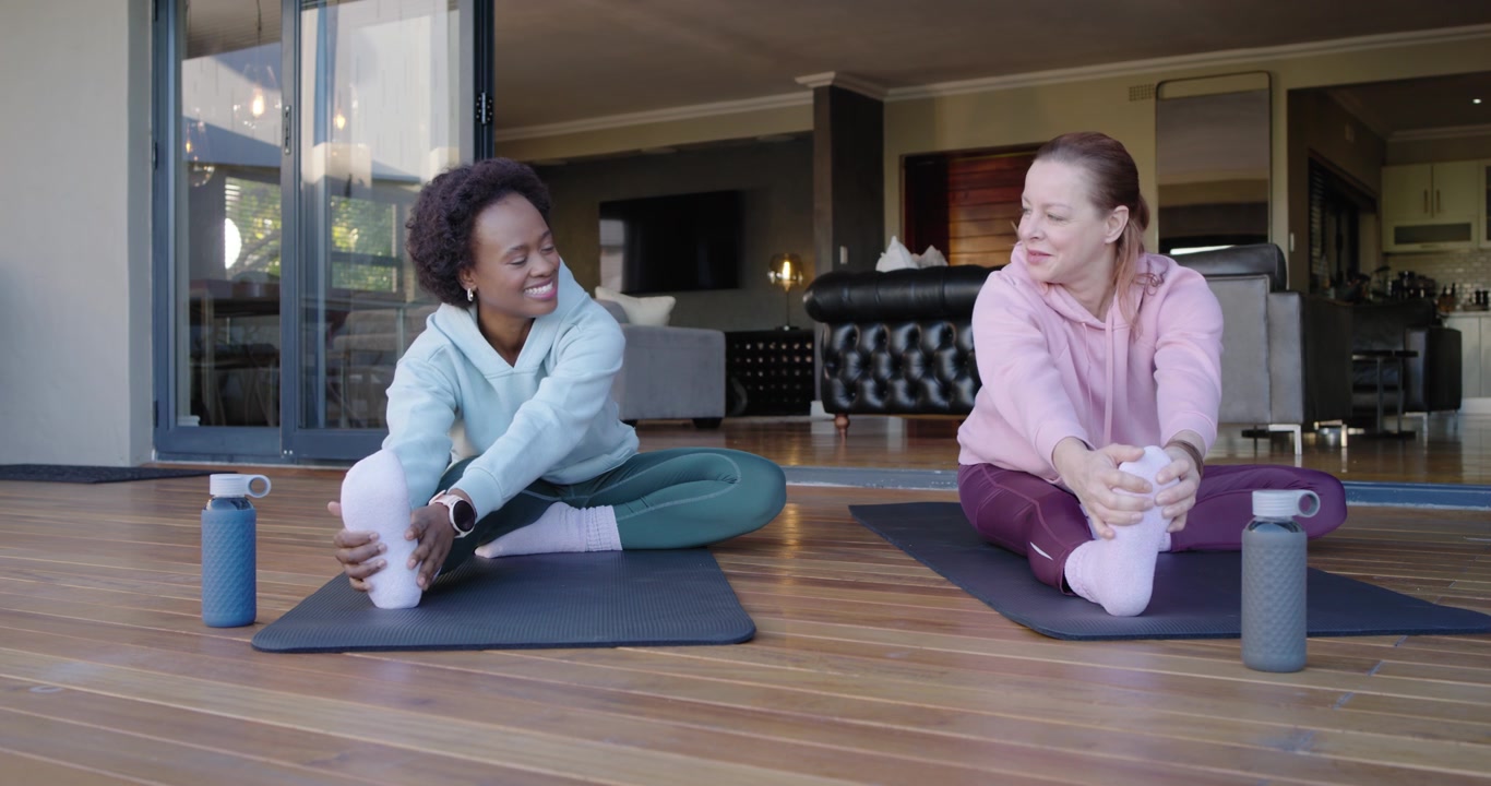 Diverse female friends starting warmup, reaching forward holding foot, rotating torsos on deck mats