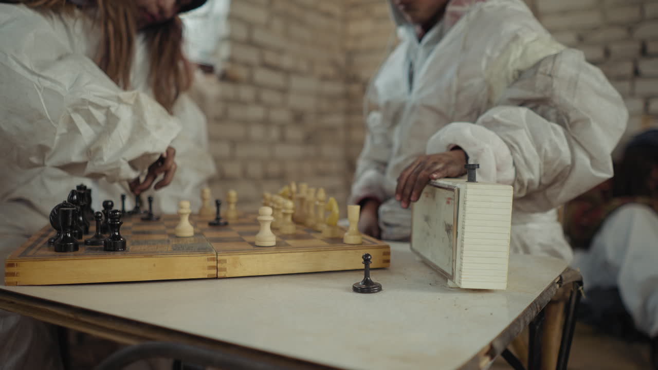 Ongoing chess game with one hand pressing timer on worn clock while opponent makes move on wooden board, both dressed in white suits inside cold room with blurred figure seated in background