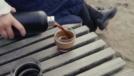 woman pouring hot coffee from thermos into mug on wooden bank at the beach in morning in slow motion