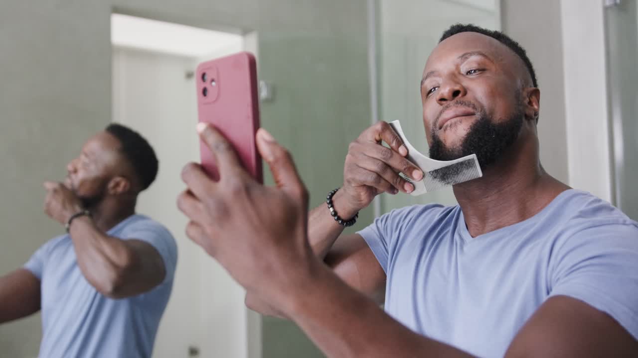 Man in 30s checking pink smartphone, smiling, pressing wipe and combing beard at bathroom mirror