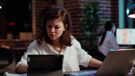 Worker-looking-over-paperwork-on-tablet,-crosschecking-with-data-on-laptop