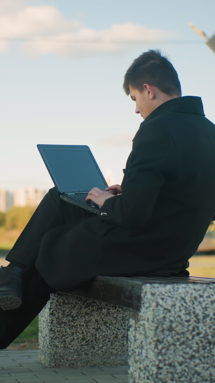 야외에 앉아 다리를 단한 채 노트북으로 작업하는 프리랜서, 초록색과 도시 환경에 둘러싸여, 배경에는 반대 차선에 주차된 자동차, 움직이는 자동차 및 현대적인 구조물이 있습니다.