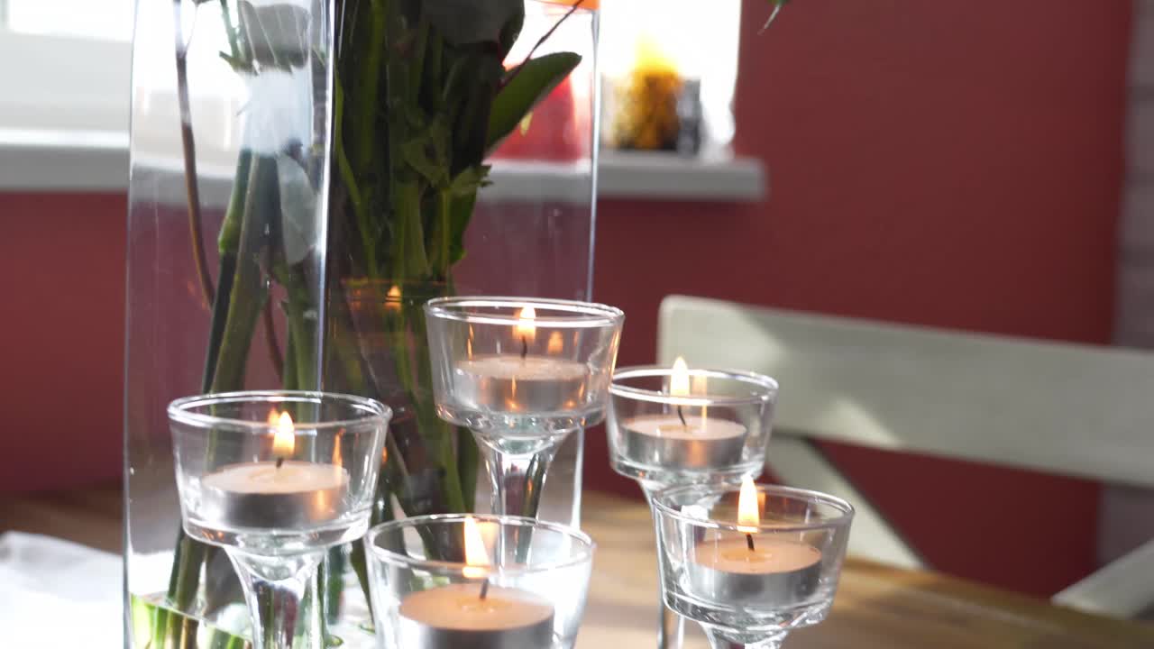 Gros plan sur un magnifique bouquet de fleurs jaunes, vertes et violettes, achetées en magasin, dans un vase en verre rectangulaire rempli d'eau. Des bougies allumées sont posées à côté, sur une table près d'un rebord de fenêtre ensoleillé. Mouvement de caméra fluide vers le bas.
