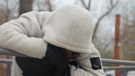 hombre cansado en suéter blanco descansando en una barra de hierro, respirando pesadamente en invierno, aliento frío visible con vista borrosa del coche que pasa en un parque urbano lejano durante una sesión de entrenamiento intenso