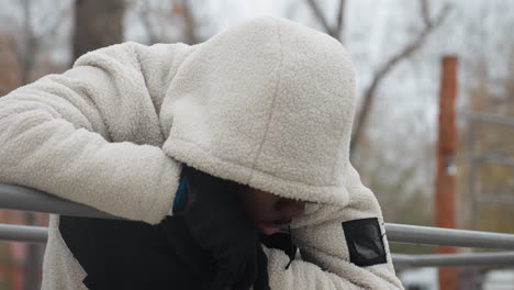 tired man in white sweater resting on iron bar, breathing heavily in winter, visible cold breath with blurred view of car passing by in distant urban park during intense workout session