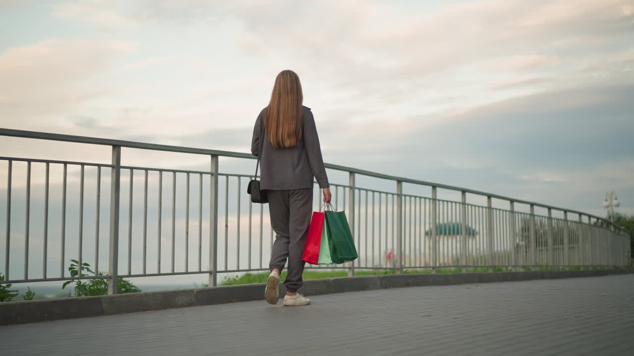 회색 옷을 입은 여자가 철 철로를 따라 고 쇼핑 가방과 검은 드백을 들고, 조용한 야외 설정과 부드러운 자연 빛 아래 배경에서 흐릿한 사람들을 가지고 있습니다.
