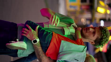 Vertical-video:-A-dad-with-curly-hair-in-a-green-T-shirt-consoles-his-little-son,-a-blond-boy-with-blue-hair,-while-they-choose-a-ticket-at-the-ticket-office-in-an-amusement-park-in-the-evening