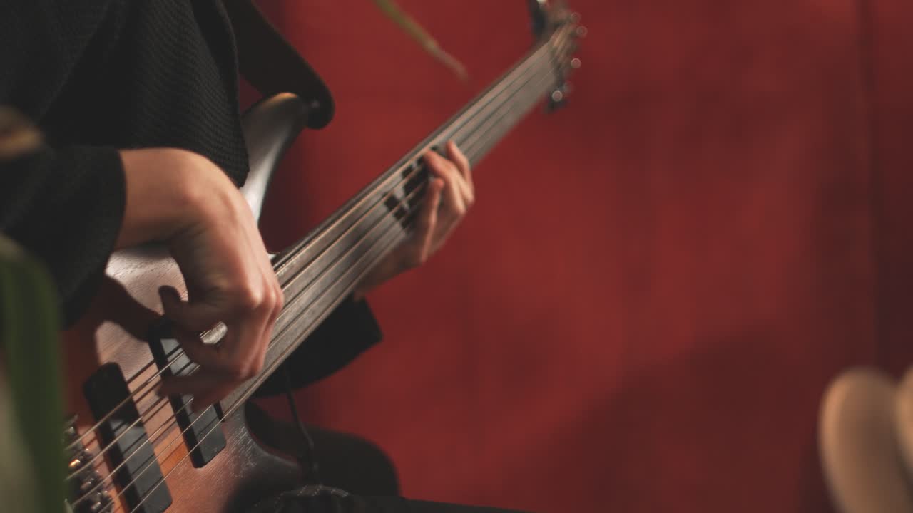 Primo piano di un musicista professionista che suona un basso elettrico fretless durante una sessione di registrazione in uno studio con una parete rossa sullo sfondo sfocato. Ripreso in 4K.