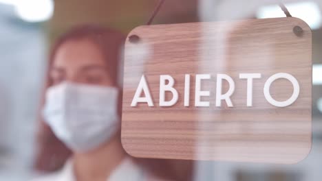 young sad woman with face mask changing open to closed sign in spanish on window for corona virus pandemic. 4k 60fps