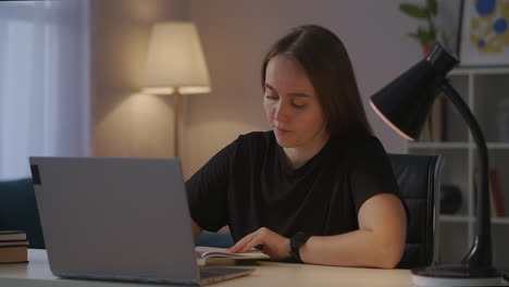 Una-Mujer-Está-Tomando-Notas-En-Un-Cuaderno-Y-Viendo-Un-Video-Tutorial-De-Aprendizaje-En-La-Pantalla-De-Una-Computadora-Portátil-Estudiando-En-Línea-En-Casa-Por-La-Noche,-Retrato-Medio