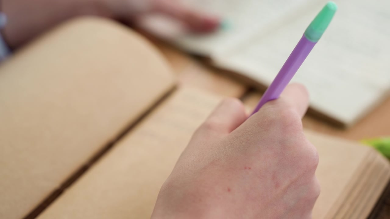 Close up of hand holding purple pen over open notebook with another person blurred hands visible in background. scene shows soft lighting and natural skin texture details focusing