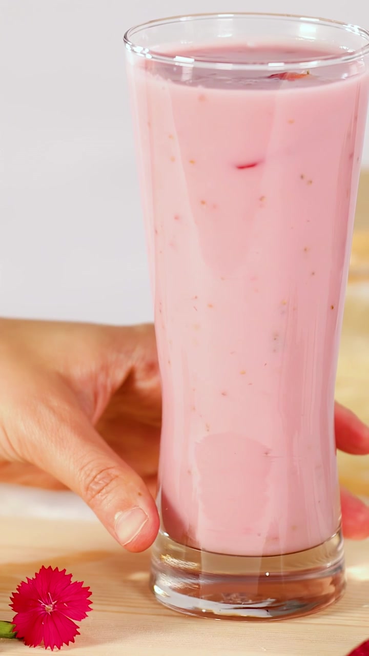 Una mano appoggia un bicchiere alto di frullato cremoso rosa alla fragola su una superficie di legno chiara.