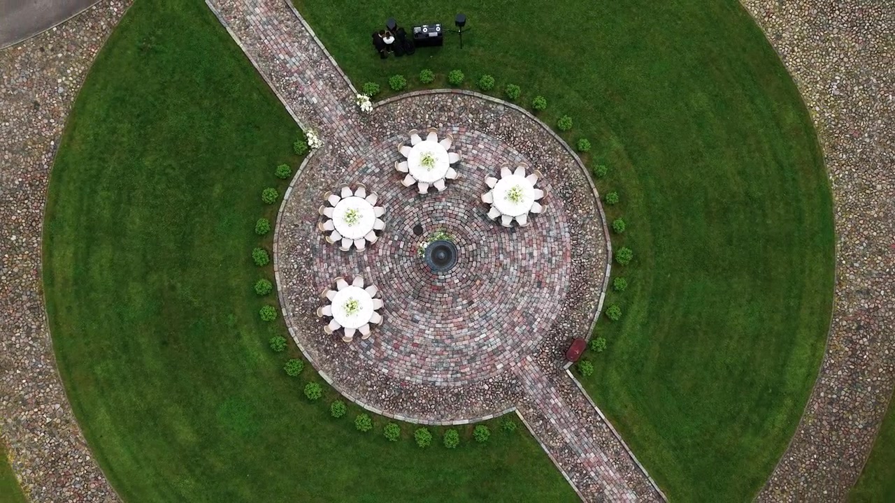 Vídeo de drone captura uma praça de jardim circular com mesas decorativas e fonte central, delimitada por gramados verdes exuberantes e caminhos de pedra, sob a luz suave do dia nublado, revelando uma simetria cuidadosa.