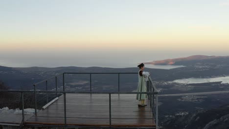 mujer con vestido tradicional montenegrino disfrutando de una copa de vino tinto parada en una terraza de madera con vista de pájaro de la bahía de kotor