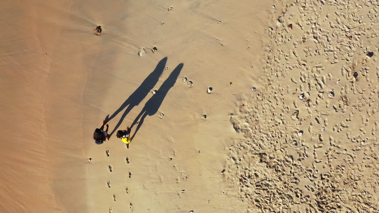 Two girls casually walking down the shore of a tropical beach, with shallow waves crashing next to them during an afternoon