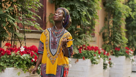 Beautiful-And-Charming-Young-Woman-In-The-Colorful-Traditional-Clothes-And-In-Headphones-Listening-To-The-Music-On-Her-Smartphone-And-Enjoying-It-In-The-Picturesque-Yard-With-Flowers